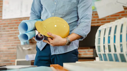 Engineer holding a hard hat and rolled technical drawings at a workshop desk